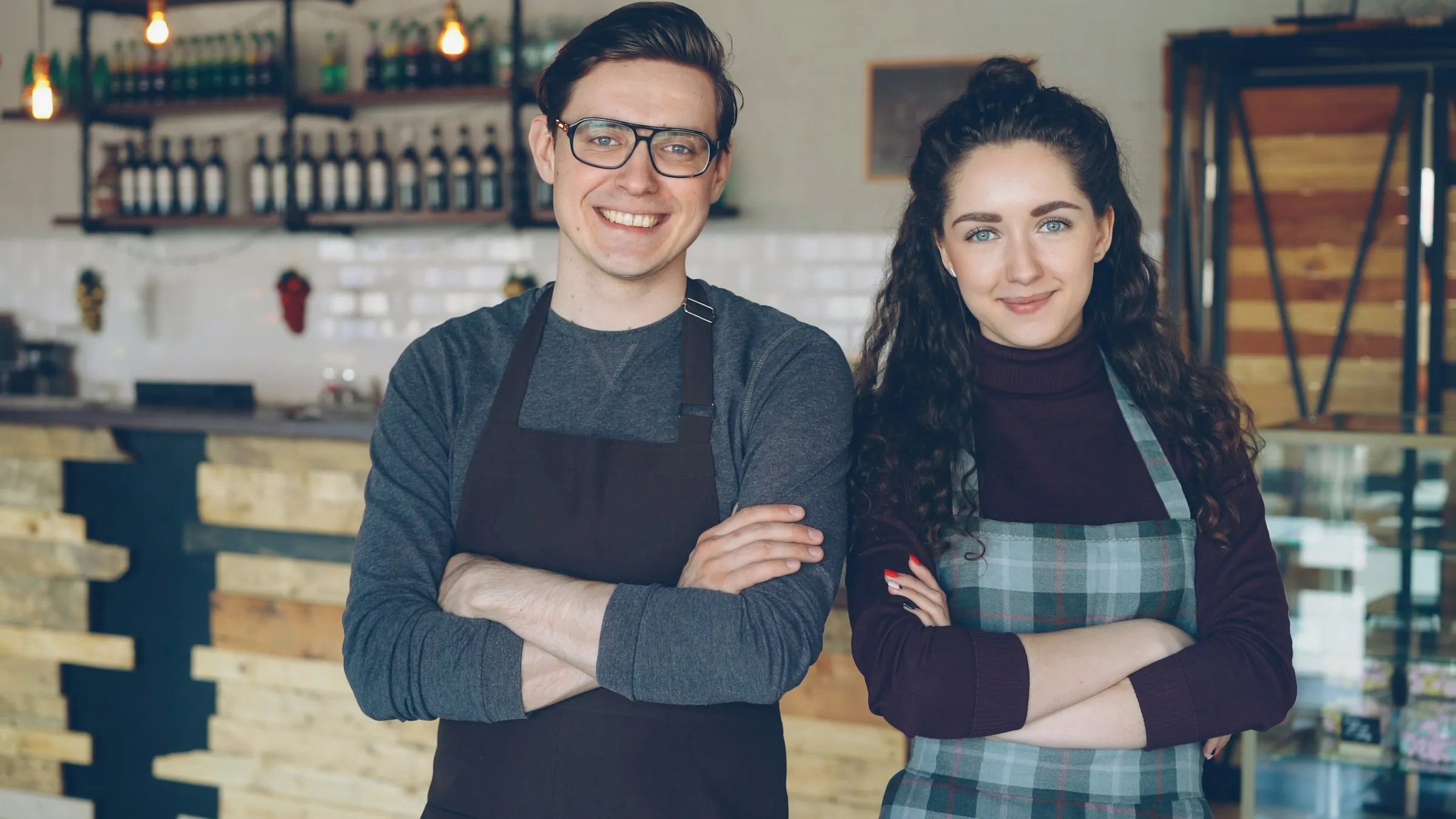 Two smiling café owners stand with arms folded in front of the counter of their café.