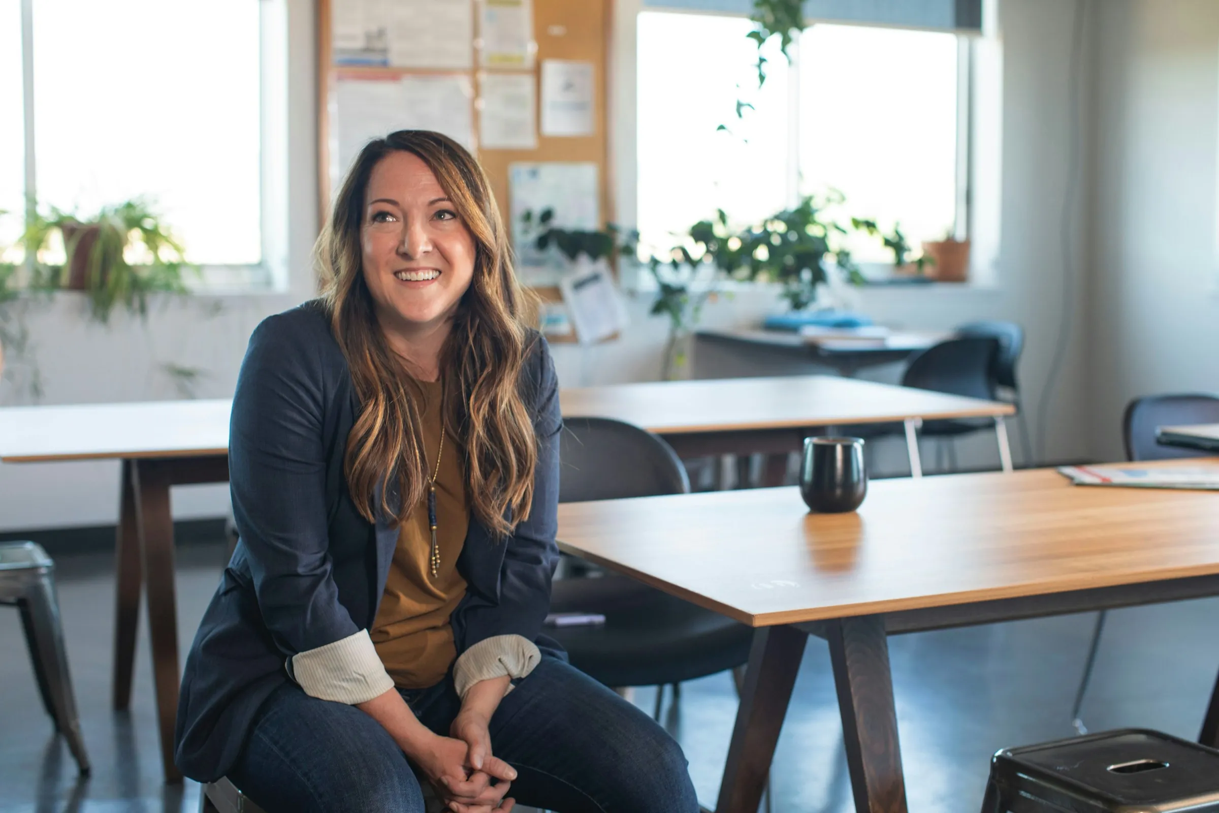 A professional in a blazer smiles as she sits at a table in a bright, plant-filled office.