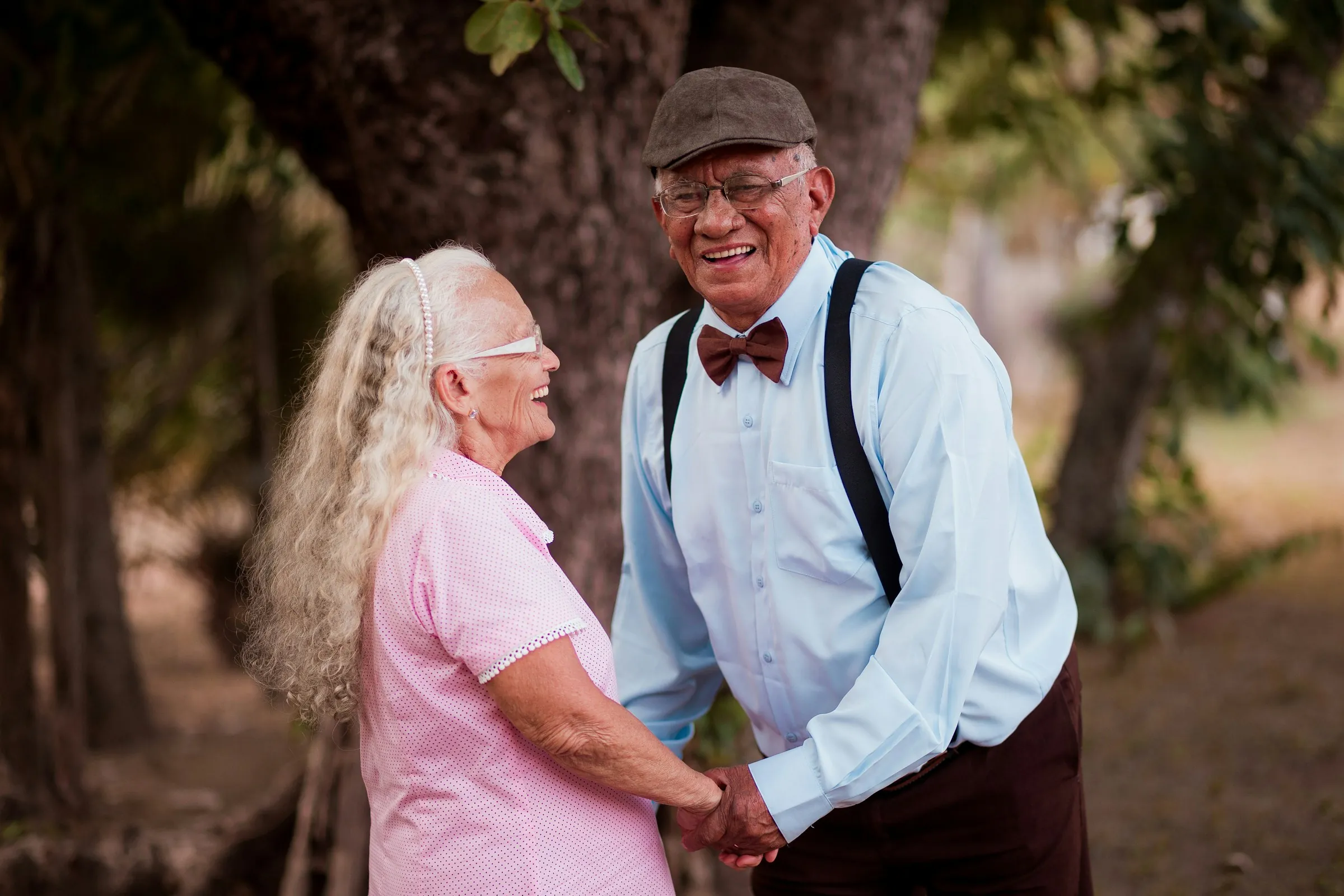 A retired couple hold hands and laugh together under the trees in a park.
